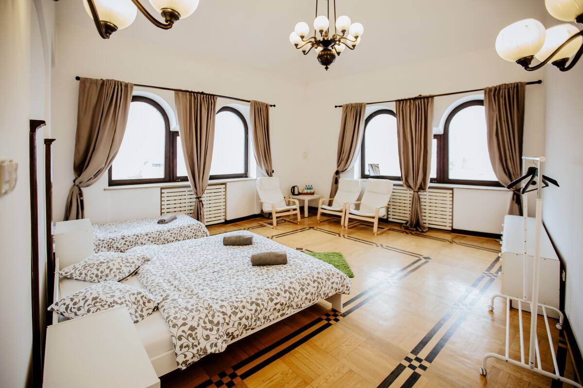 A hotel room with two beds, a desk, chairs, and large windows with curtains.