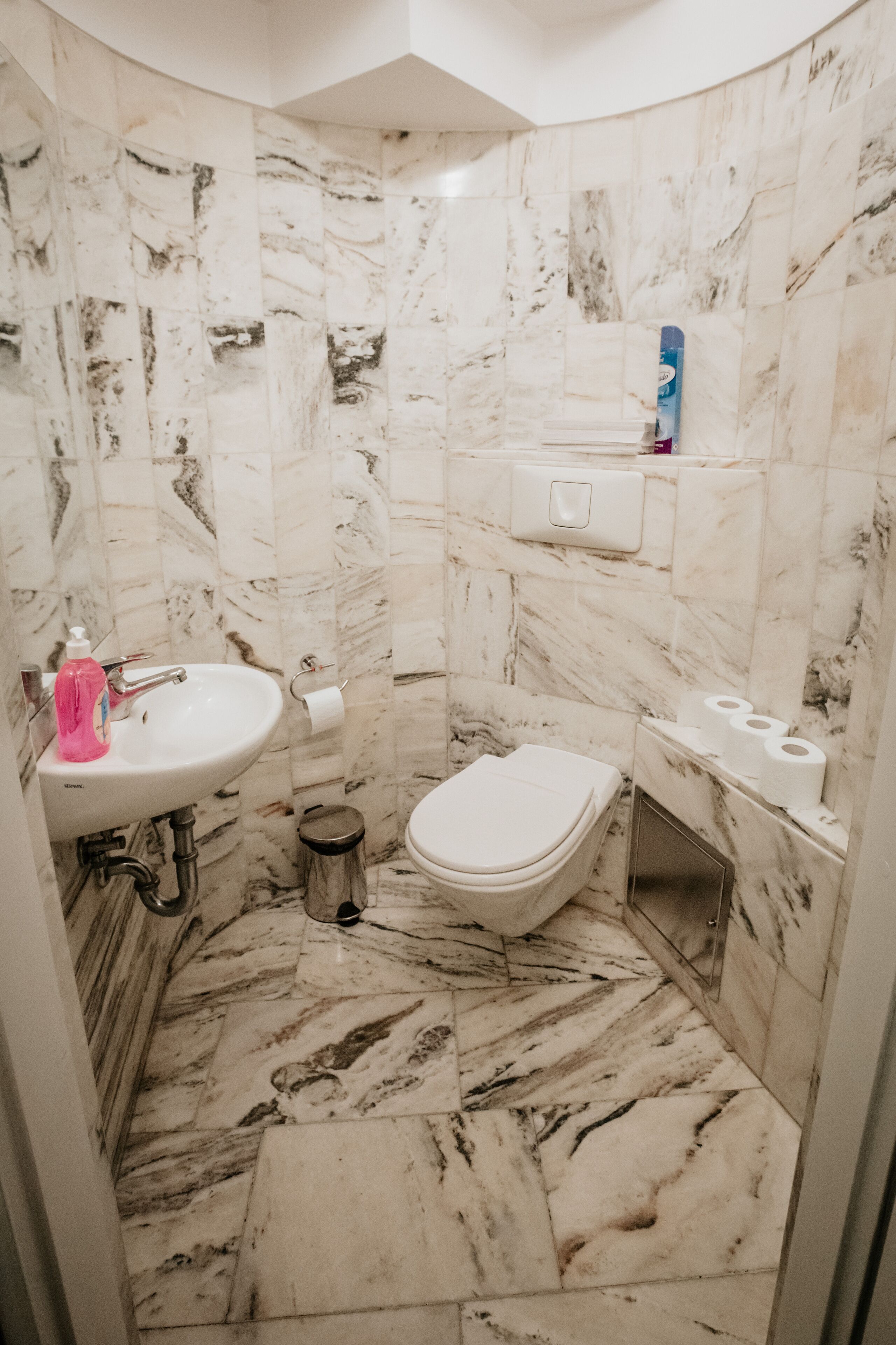 Un baño con suelo de mármol, un lavabo blanco, un inodoro y un cubo de basura.