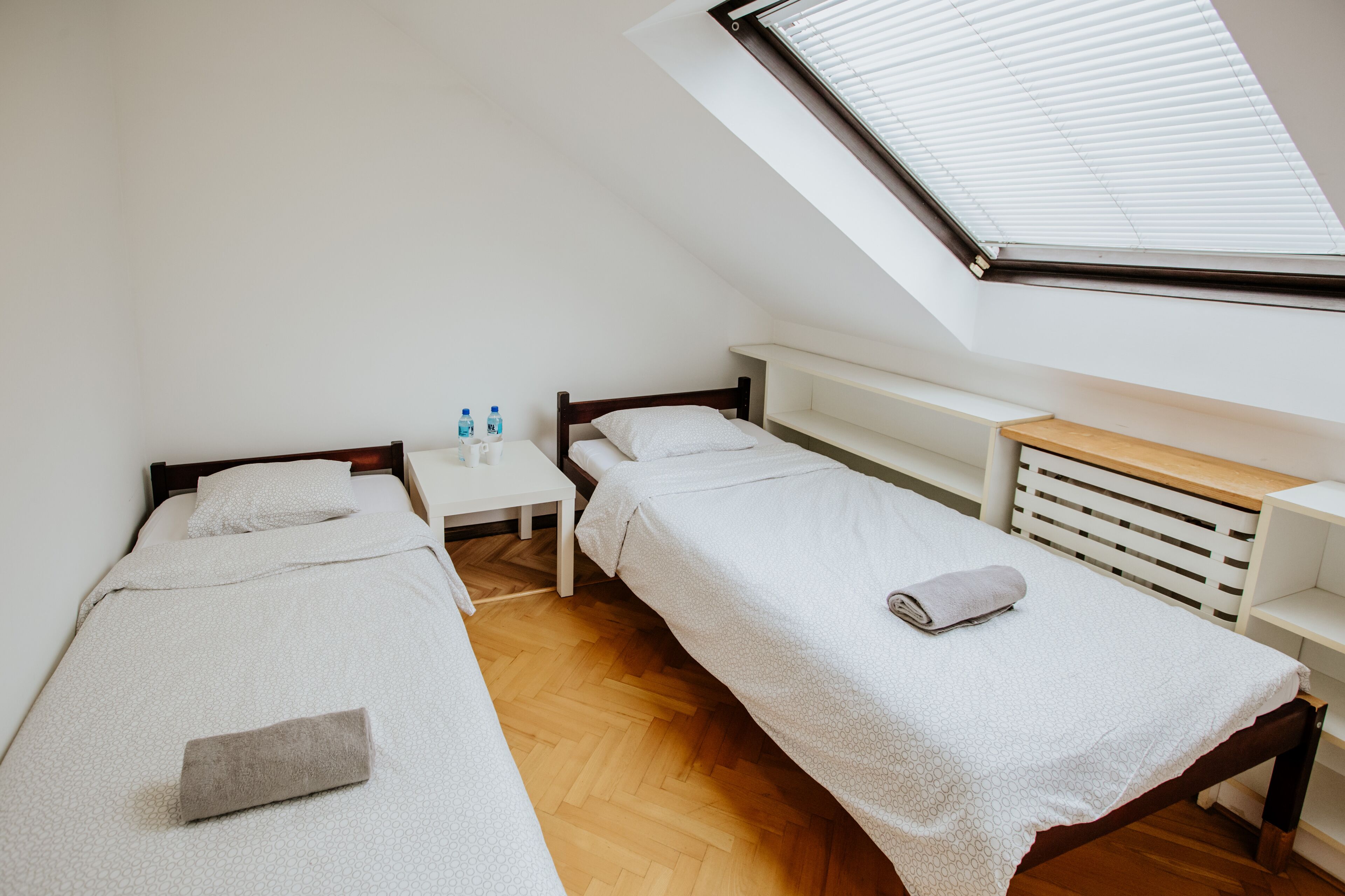 Una habitación pequeña y moderna con dos camas individuales, suelo de madera, tragaluz y una mesita de noche blanca con botellas de agua.