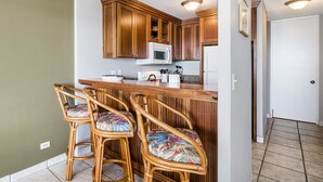 Una cocina con armarios de madera, un microondas blanco y una zona de bar con sillas de mimbre.