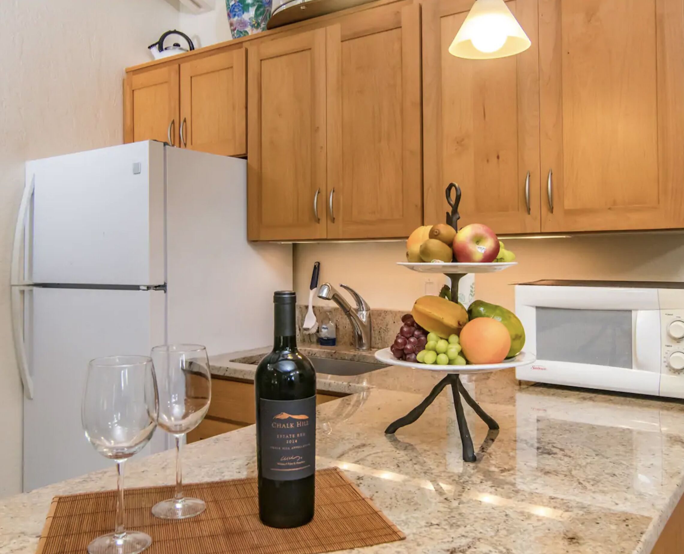 A kitchen with wooden cabinets, a white refrigerator, a microwave, and a fruit display on the counter.