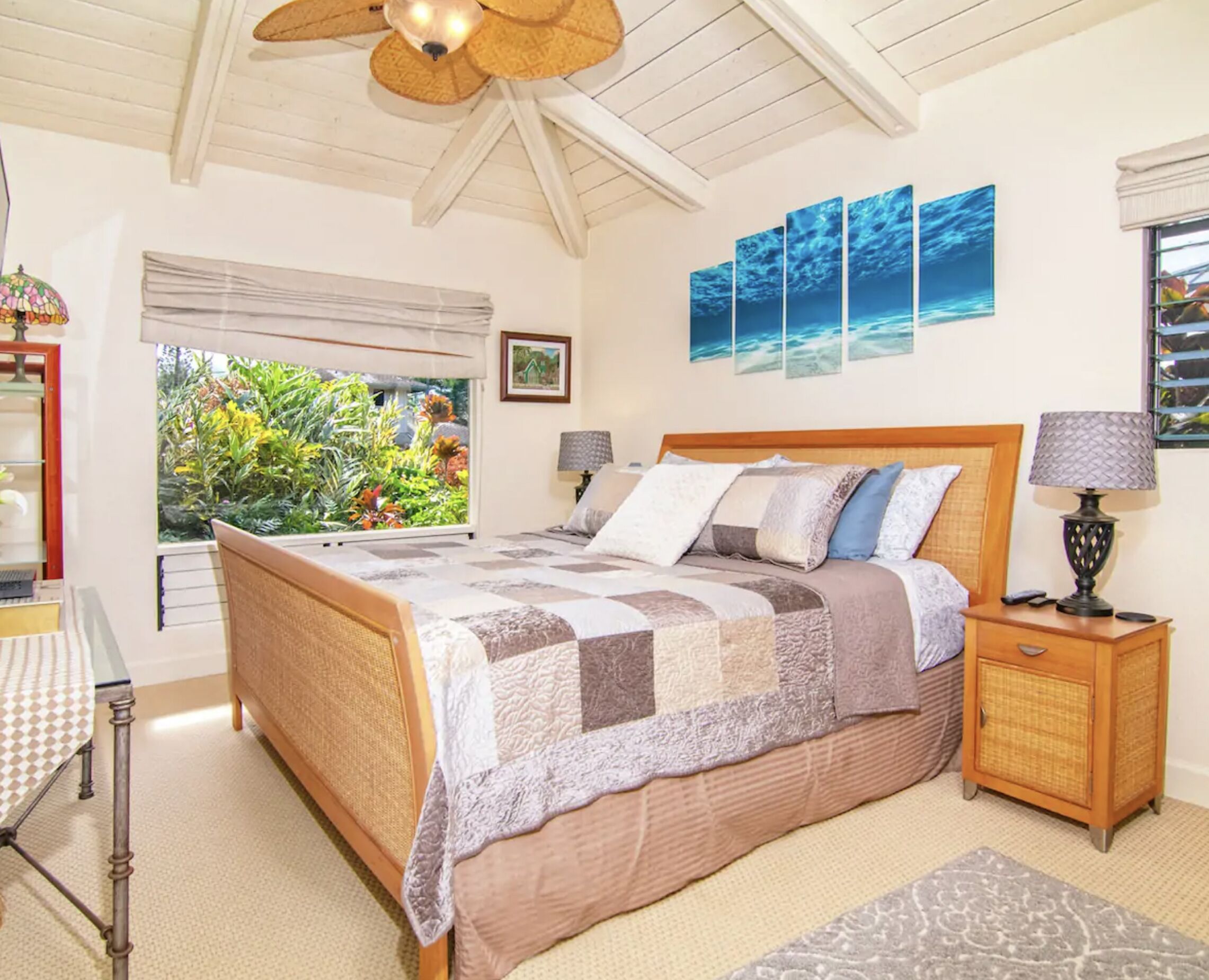 Une chambre à coucher comprenant un lit, des tables de chevet, une lampe, une fenêtre donnant sur un paysage verdoyant et un ventilateur de plafond.