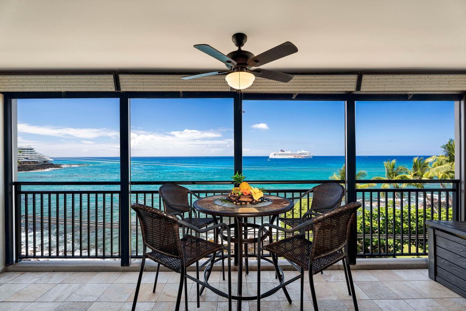 A balcony with a table and chairs, a ceiling fan, and a view of the ocean and a cruise ship.