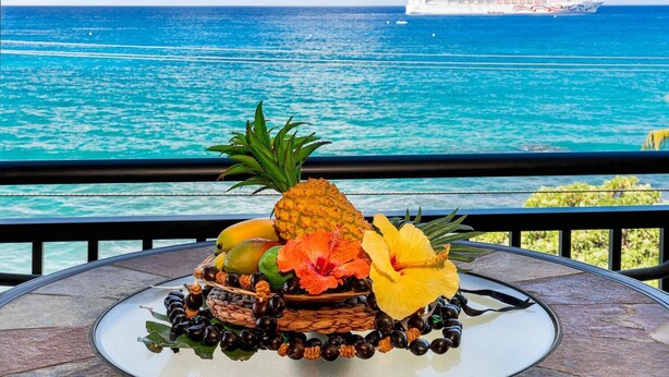 Disposición de frutas tropicales sobre una mesa, con vista a un crucero y un océano turquesa.