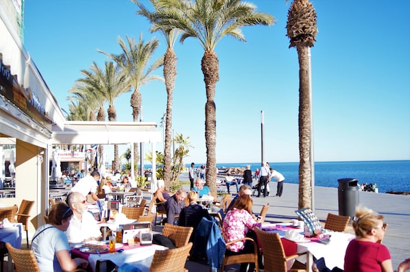 An outdoor dining area with tables and chairs, palm trees, and a view of the sea.