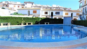 A swimming pool with a clear blue water surface, surrounded by a tiled deck. In the background, there are white buildings with terracotta roofs and multiple balconies.