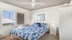 Une chambre à coucher comprenant un lit, des tables de chevet, un ventilateur de plafond, une fenêtre avec des stores et un tableau encadré accroché au mur.
