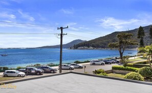 Vista sulla costa con auto parcheggiate, cielo sereno e una barca sull'acqua.