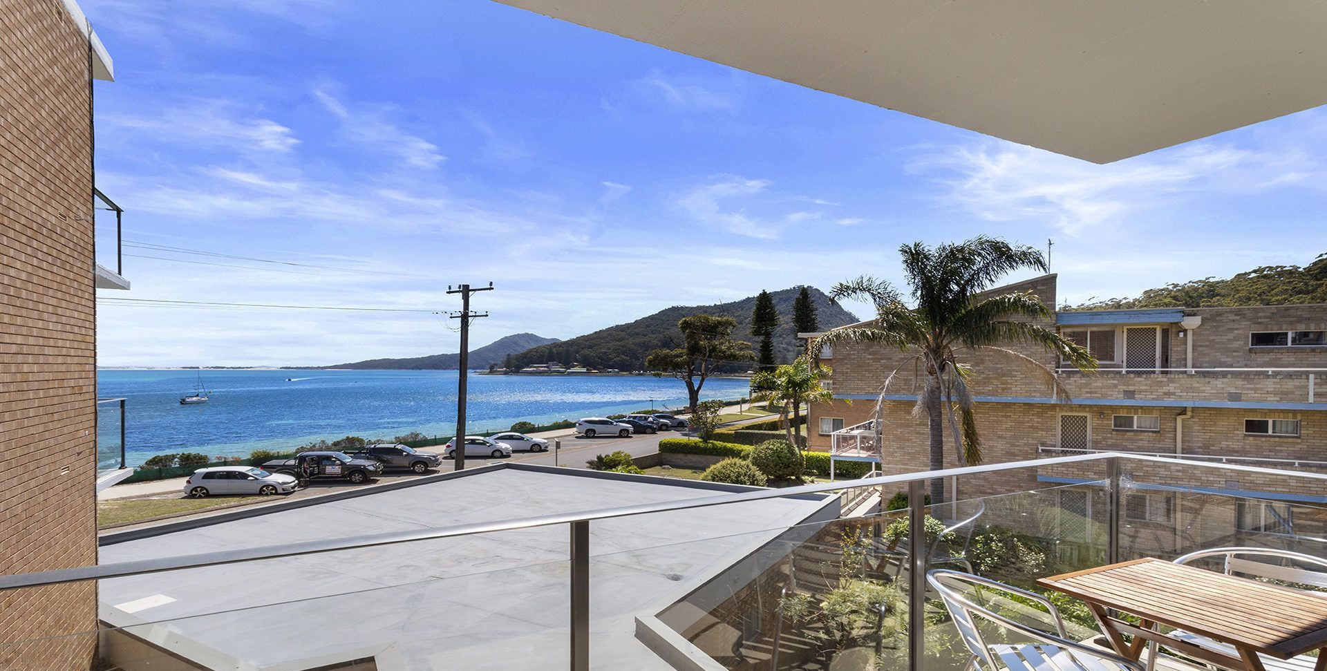 A balcony with a view of the sea, parked cars, and a building with a brick facade.