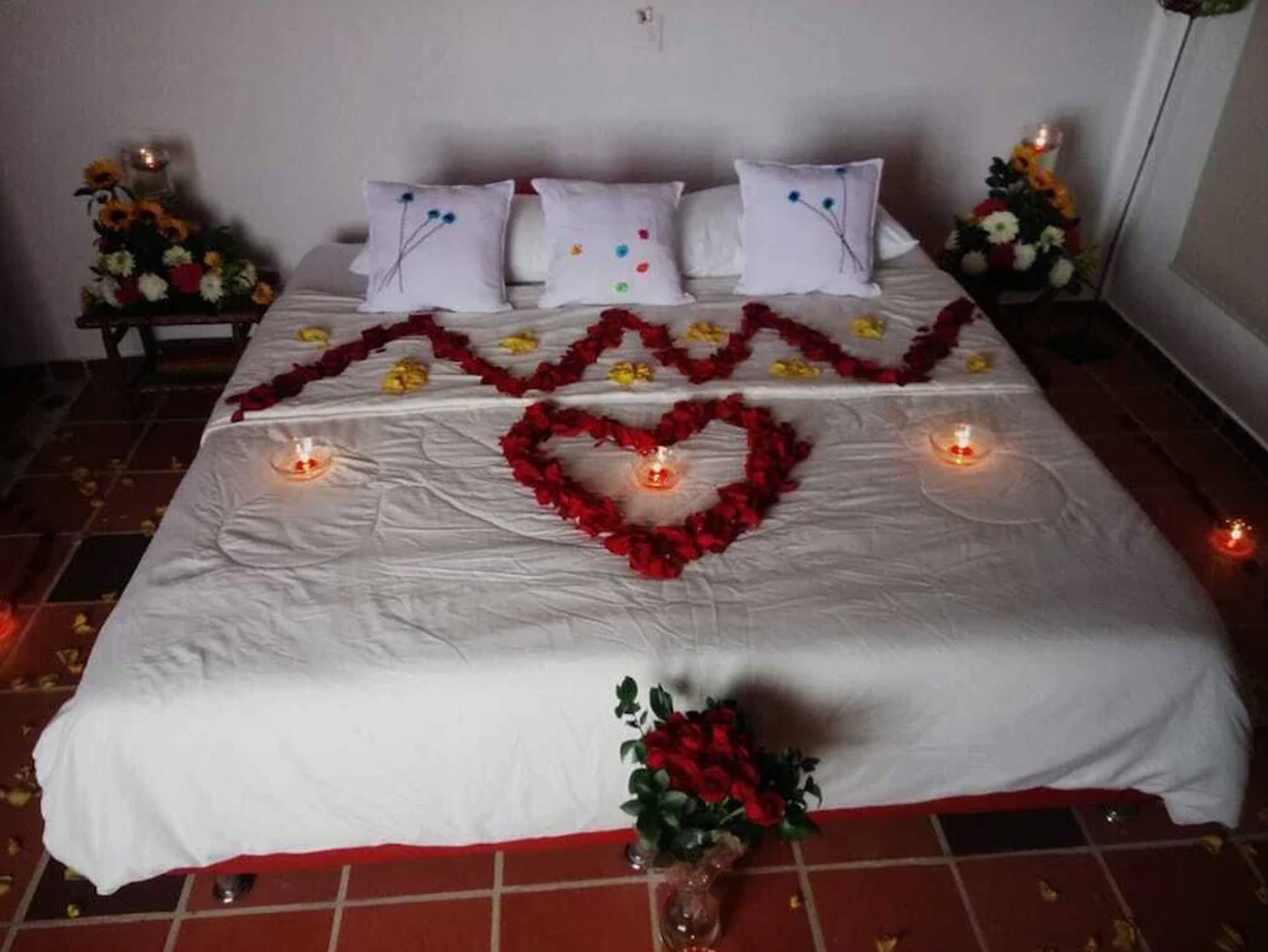 A bed with white linens, a heart-shaped decoration made of red flowers, and a bouquet of red roses at the foot of the bed.