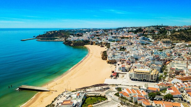 Vista aèria d'una vila costanera amb una platja de sorra, un moll i edificis densament agrupats amb arquitectures diverses.