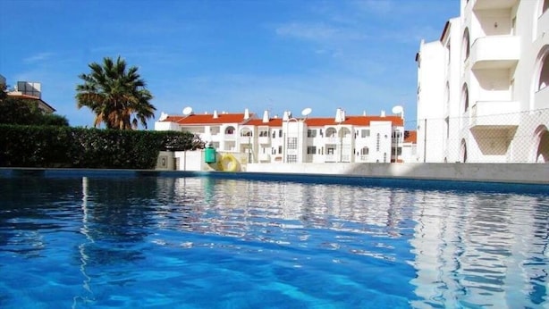 A swimming pool with clear blue water, a white building with red roofs, and a palm tree in the background. - Apartment, with pool, near the beach (Albufeira)