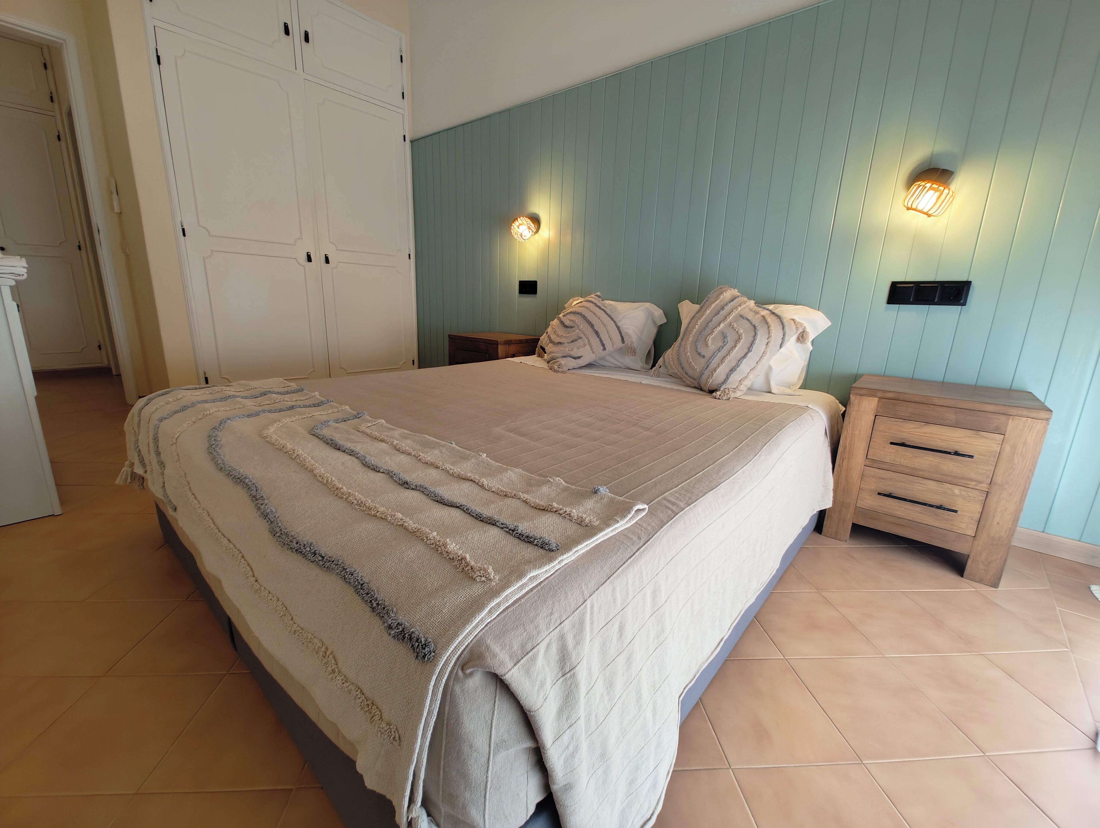 A bedroom with a large bed, two bedside tables, a wardrobe, and wall-mounted lighting.