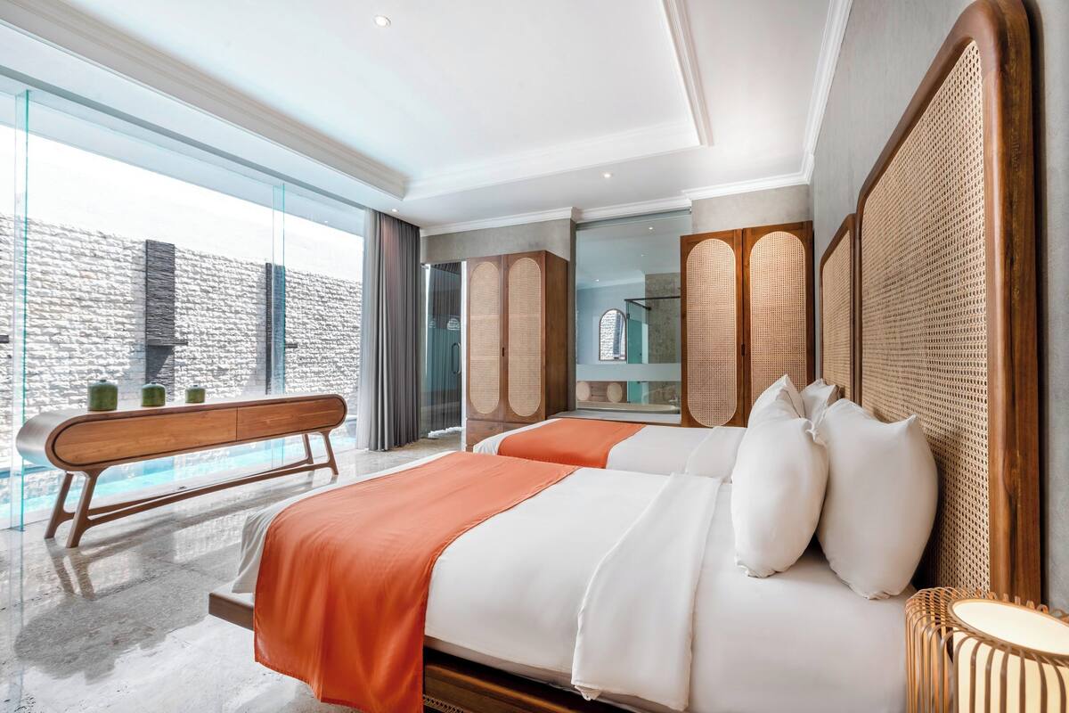 A modern hotel room with a large bed, a bench, and a view of a building through a glass wall.