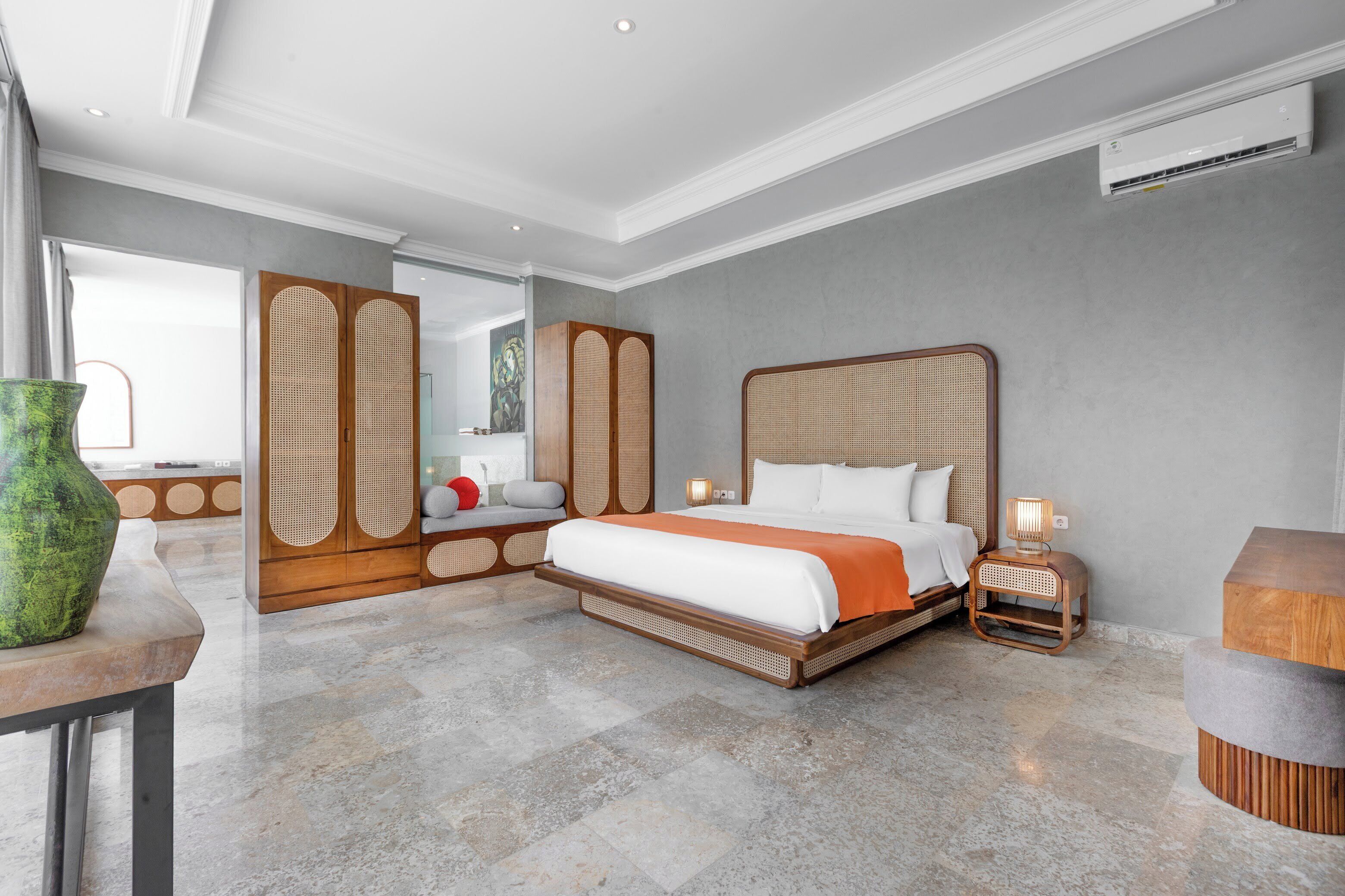 Quarto de hotel moderno com uma cama grande, um guarda-roupa de madeira, uma pequena mesa com um vaso verde e um banquinho redondo.