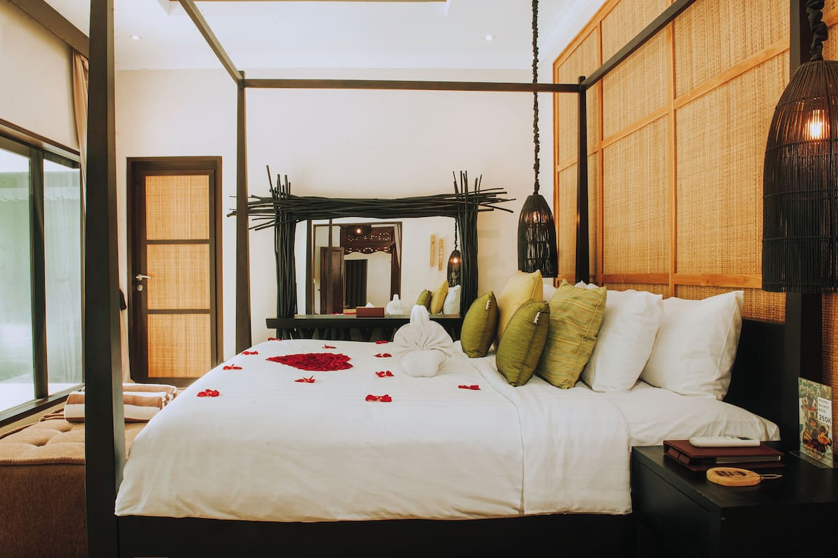 A neatly made bed with white linens and green pillows, a bedside table with a book and a phone, and a large mirror above the bed.