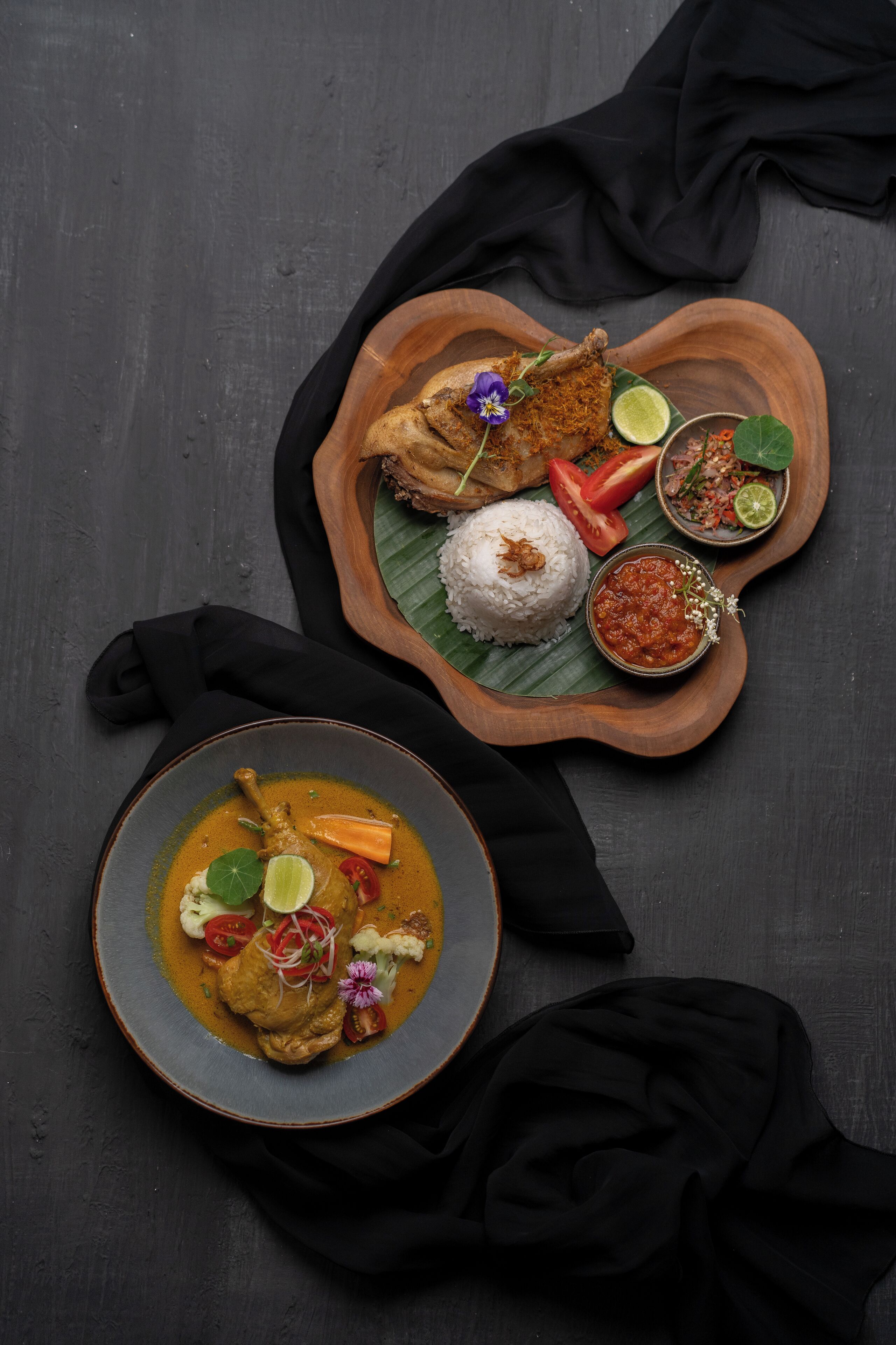 Una bandeja de madera con un plato de pollo, arroz y diversos condimentos sobre una superficie oscura.