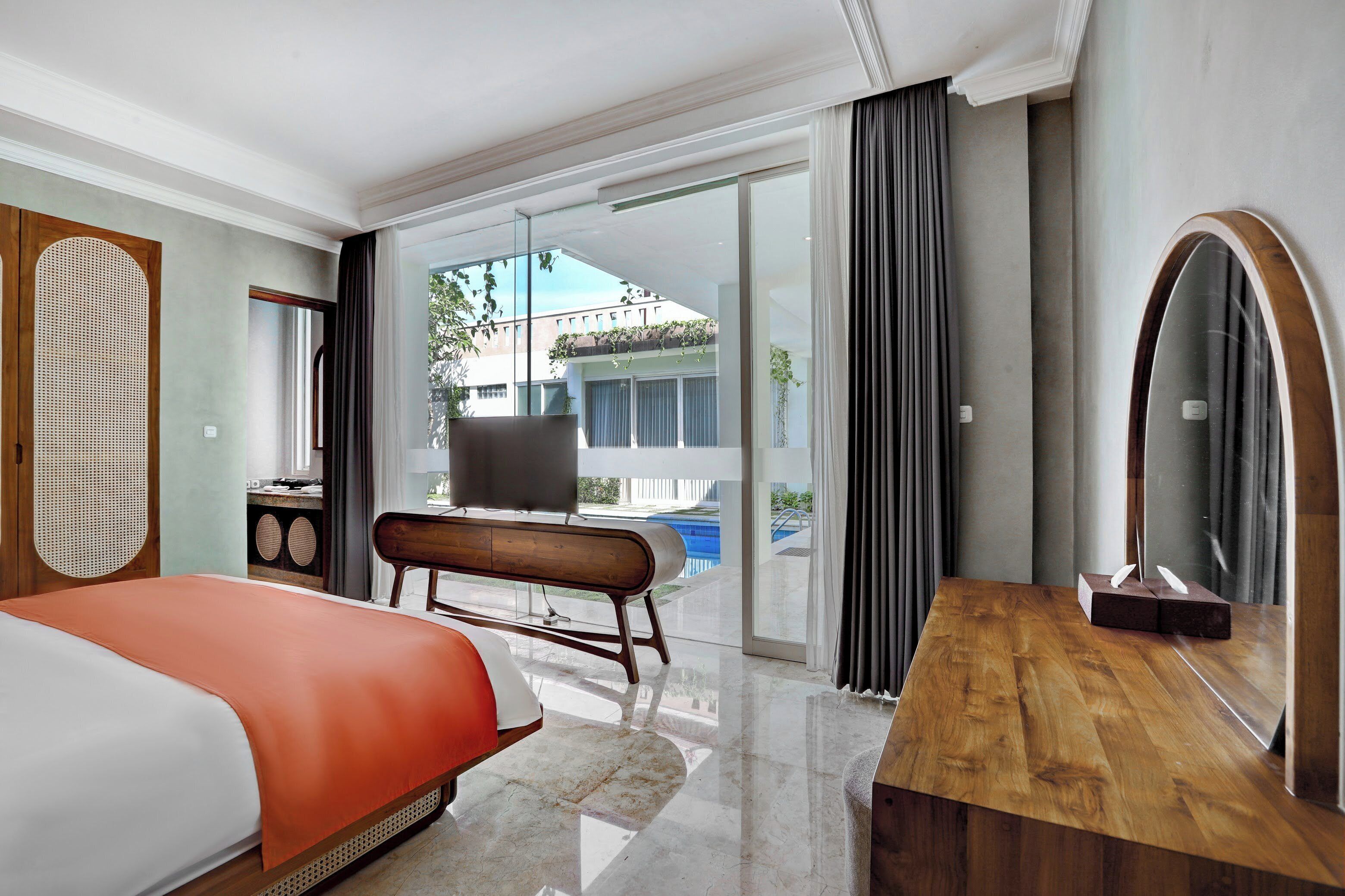 A hotel room with a bed, a wooden bench, a desk, a large mirror, and a sliding glass door leading to a balcony with a pool.
