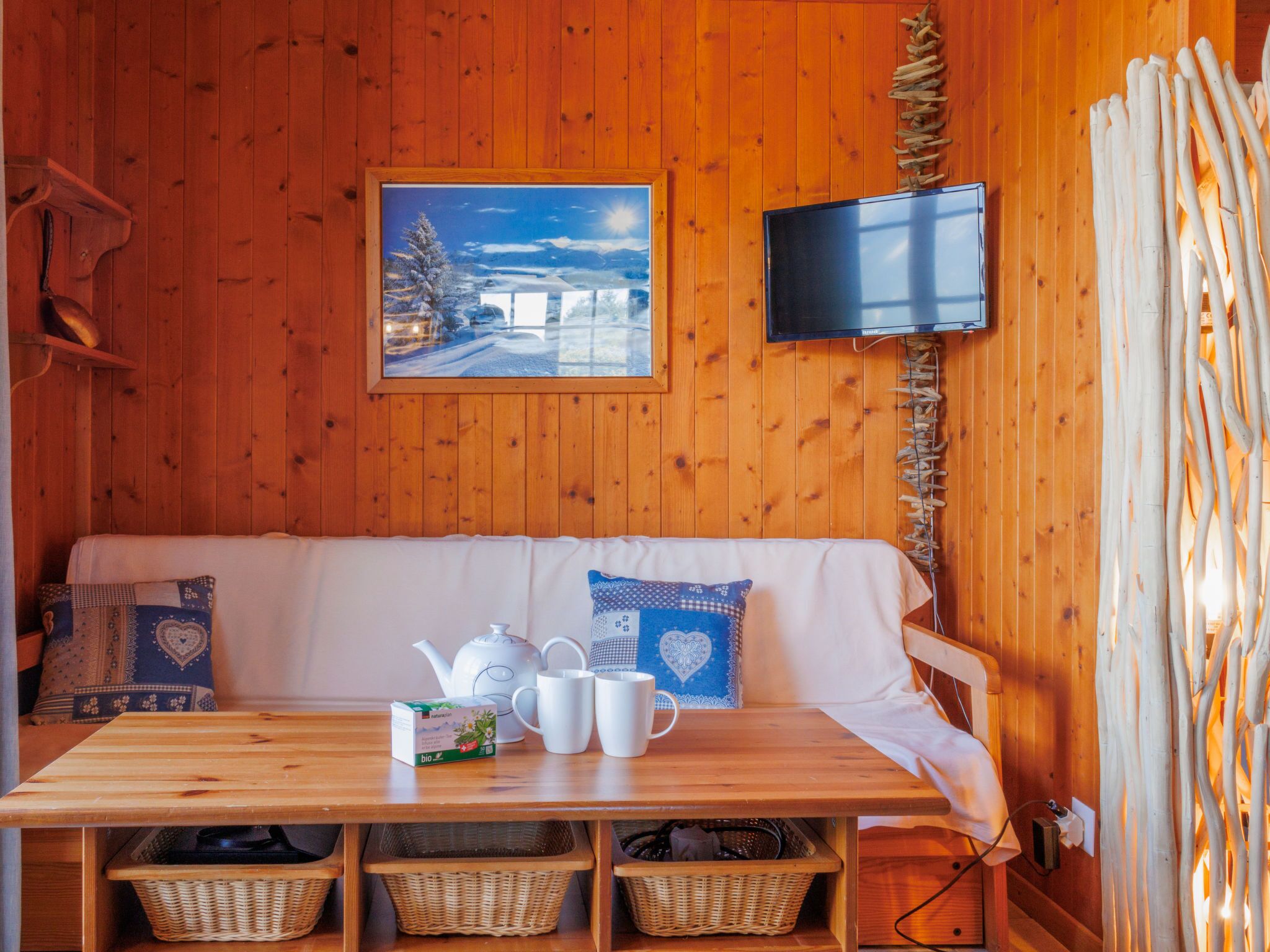 Ein gemütliches Blockhaus-Interieur mit einer hölzernen Bank, einem Tisch mit Teeservice und einem an der Wand befestigten Fernseher.