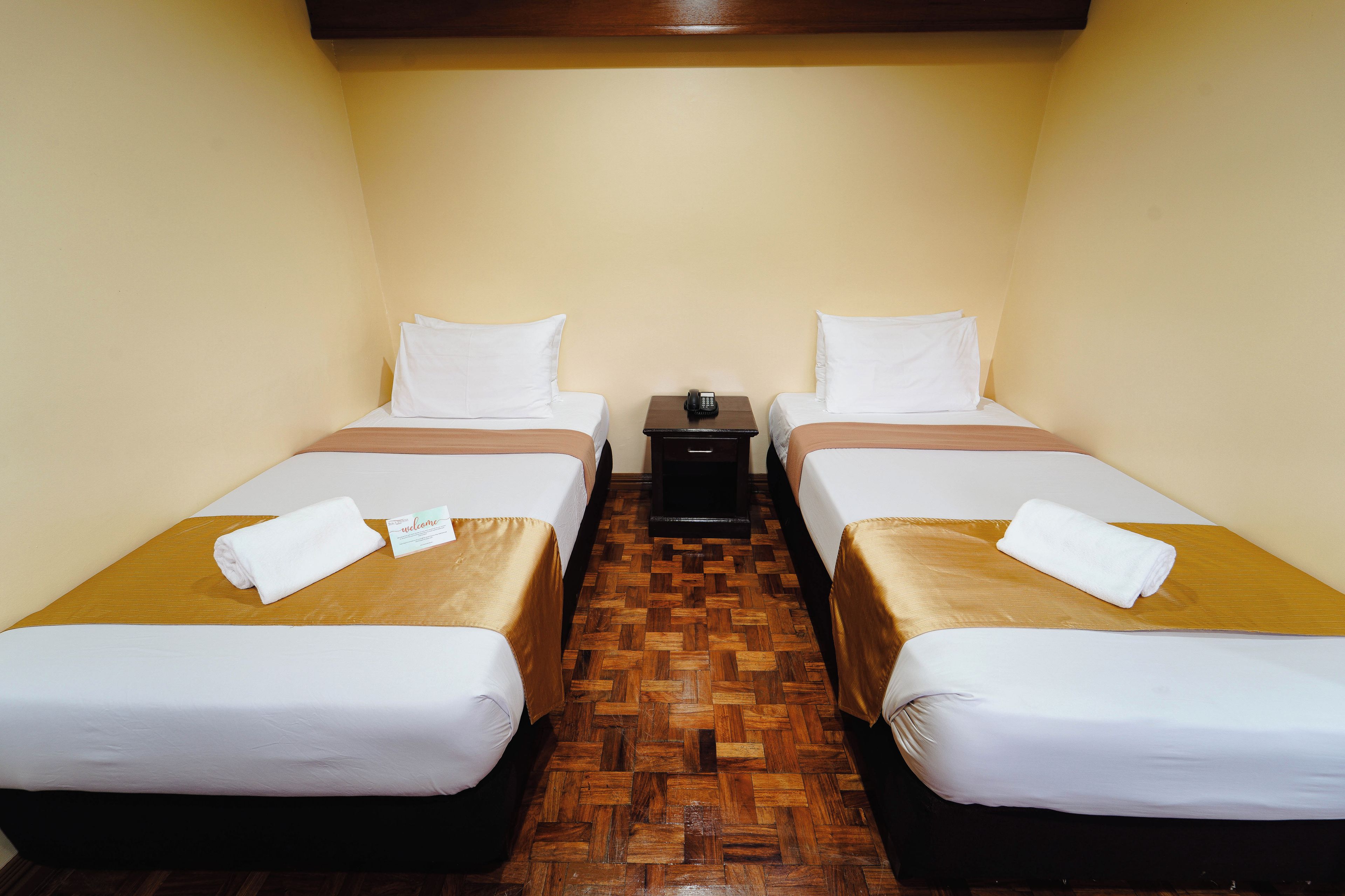 Habitación de hotel con dos camas individuales, suelo de parqué de madera y una mesita de noche con lámpara.