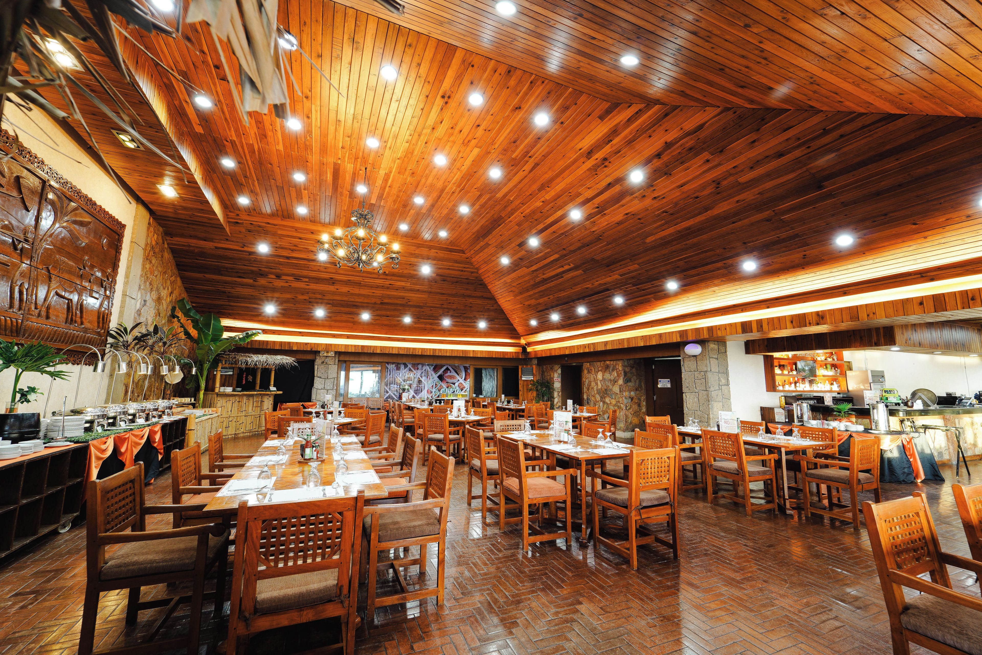 Un restaurante espacioso con suelo y techo de madera, varias mesas y sillas para comer, y una cocina visible al fondo.