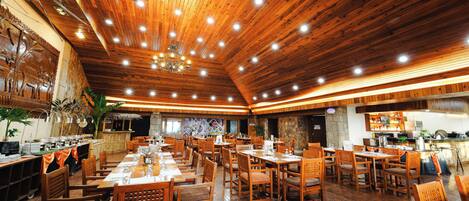 A spacious restaurant with wooden flooring and ceiling, multiple dining tables and chairs, and a kitchen area visible in the background.