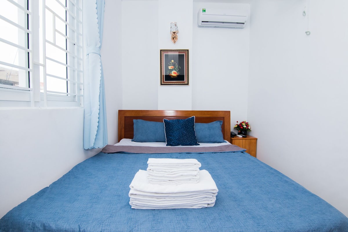 A neatly made bed with a blue bedspread and pillows, a stack of white towels, a framed picture on the wall, and a small bedside table with a vase of flowers.