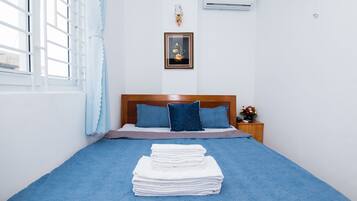 Una cama bien tendida con colcha y almohadas azules, una pila de toallas blancas, un cuadro en la pared y una mesita de noche con un florero.
