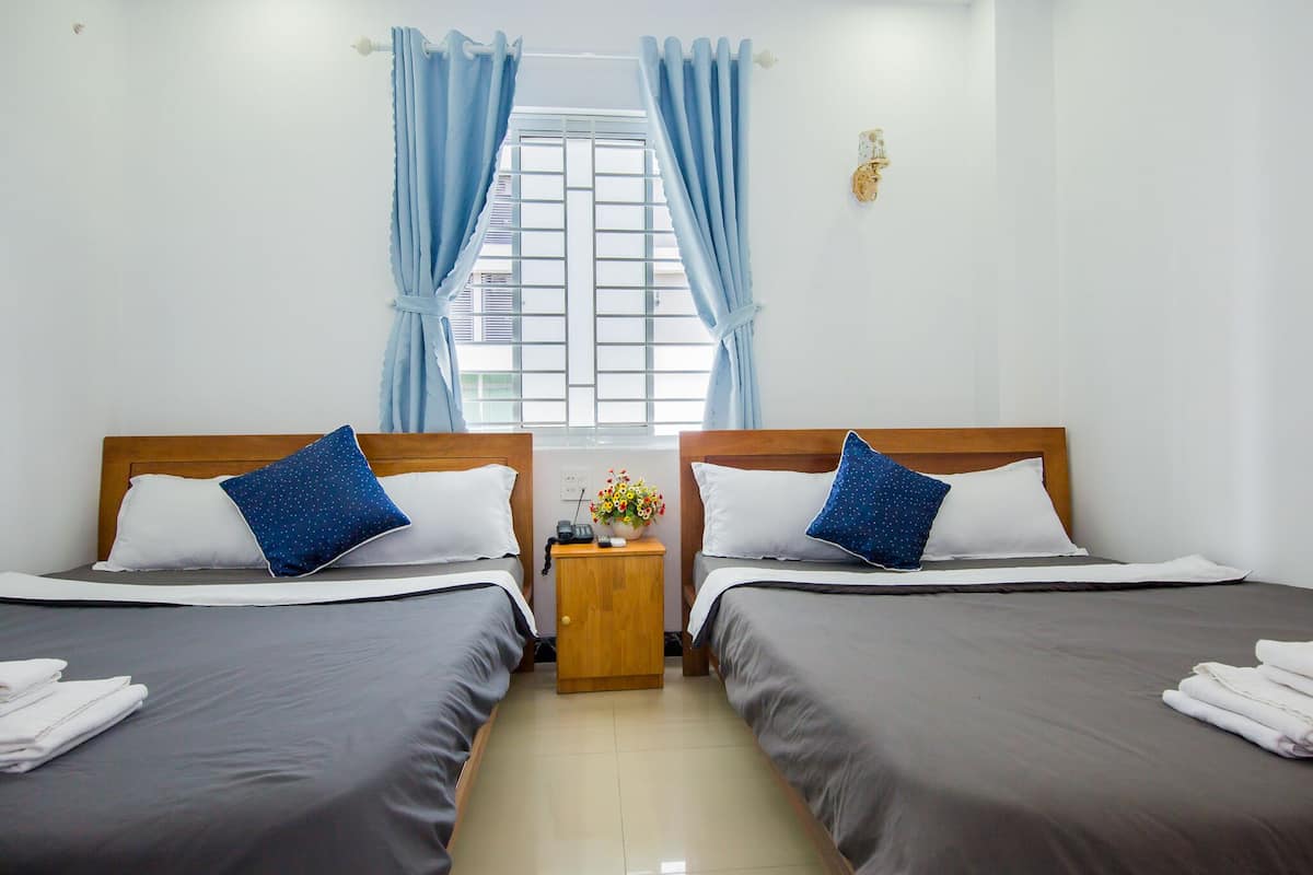 A hotel room with two beds, a wooden nightstand with a radio and a vase of flowers, and a window with blue curtains.