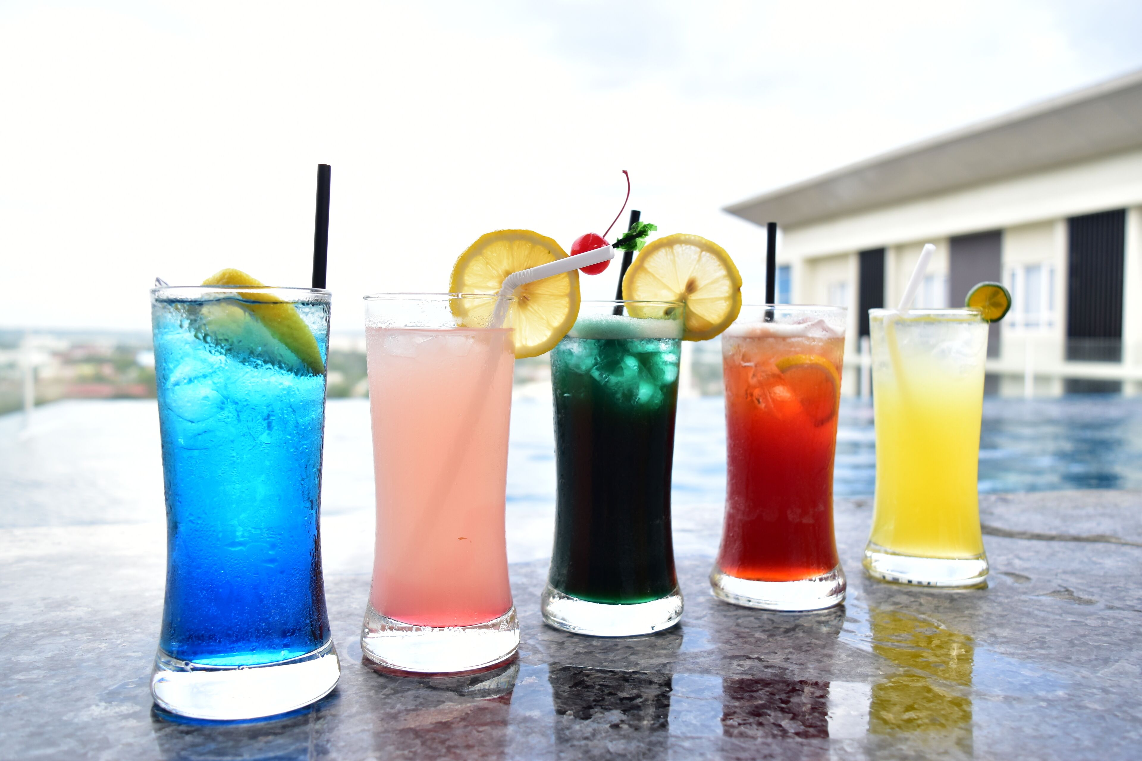 Cinq boissons colorées sont disposées sur une table, avec un bâtiment visible en arrière-plan.