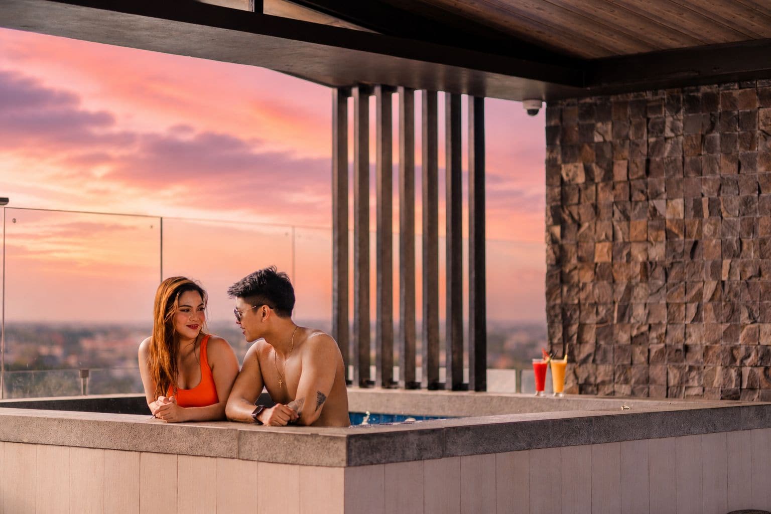 Una pareja en un jacuzzi con vista al atardecer, pared de piedra y columnas verticales.