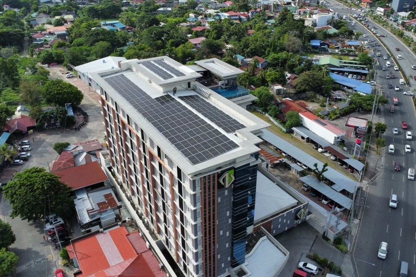Een modern gebouw met zonnepanelen op het dak, omringd door een woongebied met verschillende gebouwen en geparkeerde auto's.