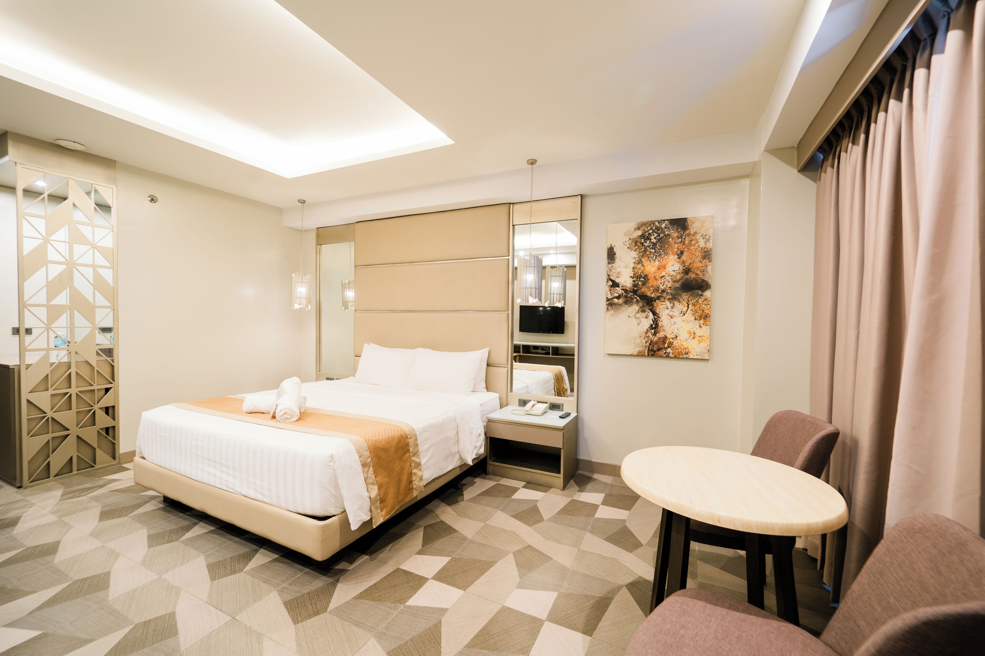 A modern hotel room with a large bed, a bedside table, a desk, and a small round table.