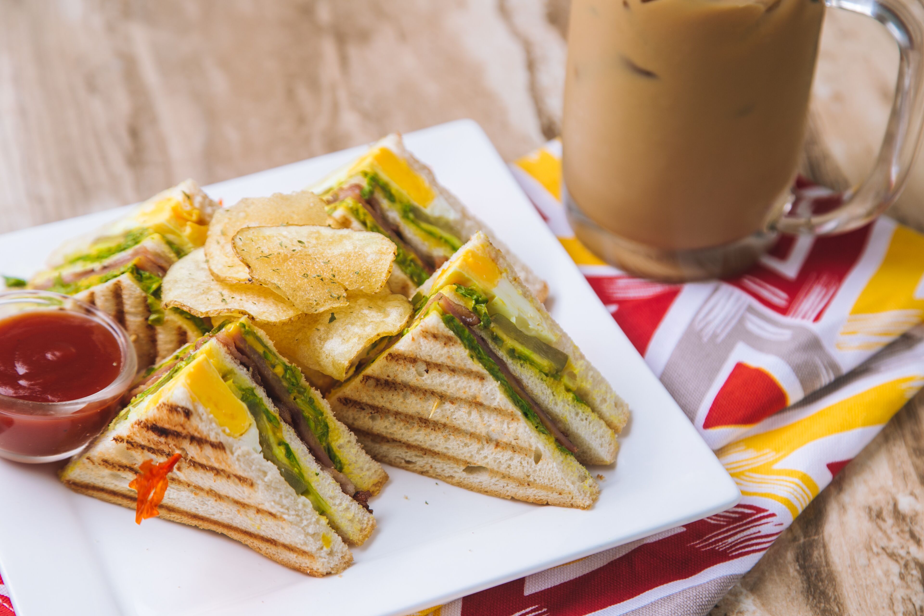 Un plato de sándwiches a la parrilla, papas fritas y kétchup, junto con una taza de café sobre una mesa con servilleta a cuadros rojos y blancos.