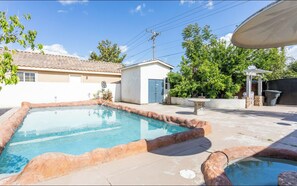 A swimming pool with a stone border, a small white building, and a pergola with a roof.