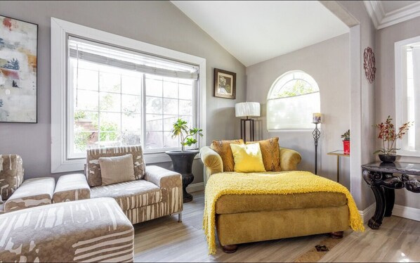 A well-lit room with a large window, a bed with a yellow blanket, a sofa, and a side table with a lamp.