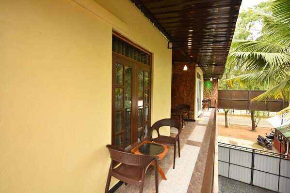 A covered outdoor seating area with wicker chairs and a table, a palm tree, and a view of the street. - The Summer Resort (Udawalawa)