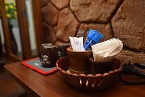 A wooden table with a red placemat, a ceramic pitcher, a wooden bowl containing napkins and a phone, and a wall with a textured pattern. - The Summer Resort (Udawalawa)