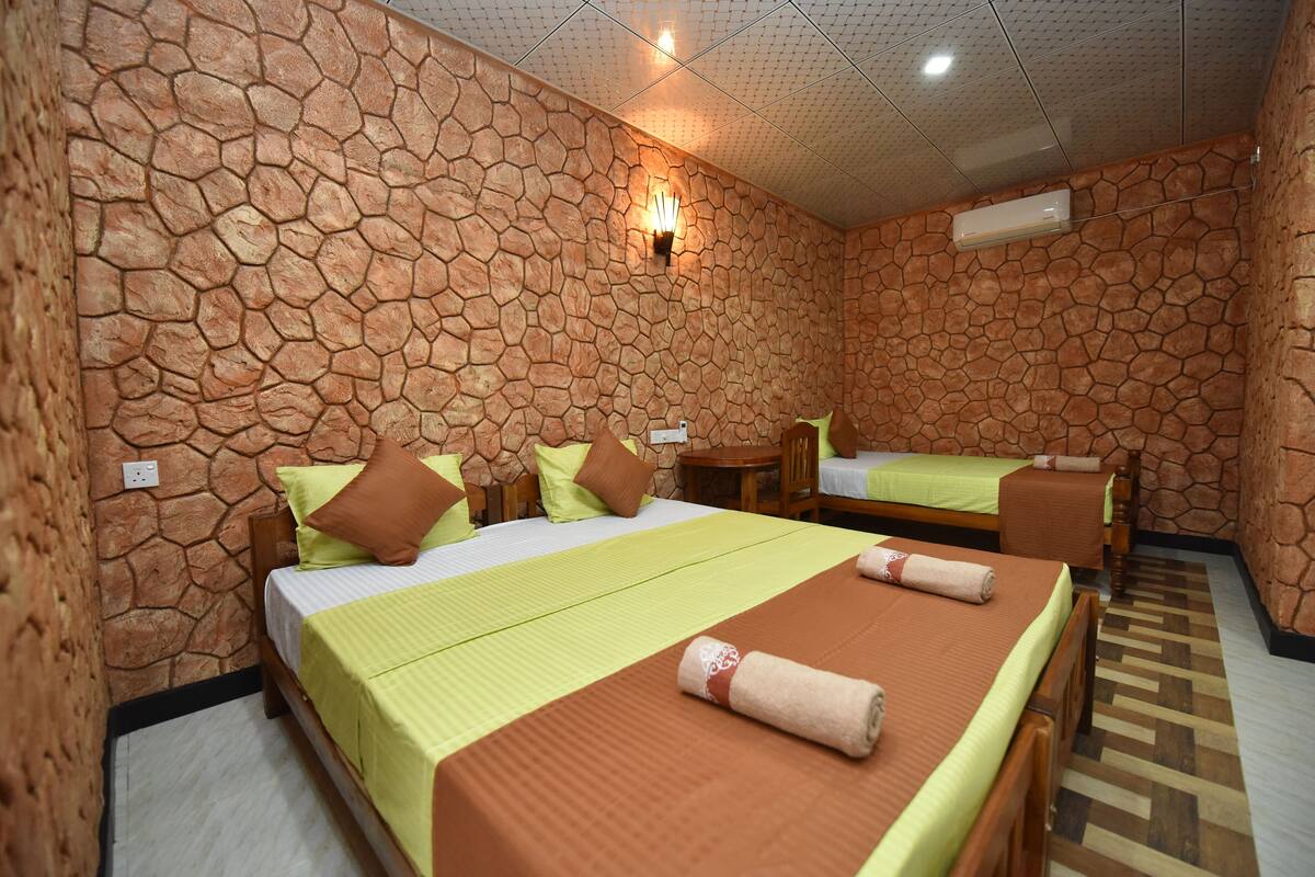 A hotel room with a stone wall design, two beds with green and brown bedding, a wooden chair, and a small table.