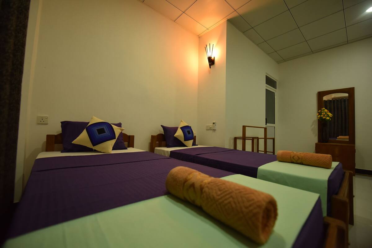 A hotel room with three beds, each with purple bedding and blue pillows. There is a wall-mounted light fixture and a mirror with a flower arrangement.