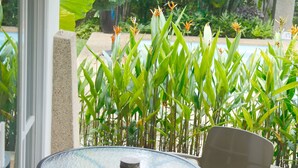 A patio with a round table and two chairs, surrounded by lush greenery and a view of a pool.