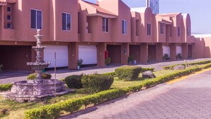 A row of modern, multi-story buildings with uniform architecture, featuring a central courtyard with a fountain, neatly trimmed hedges, and a paved walkway. - Motel LA Autoparador (Morelia)