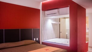 A modern bedroom with a large bed, a red accent wall, and a built-in window with a view of the bathroom. - Motel LA Autoparador (Morelia)