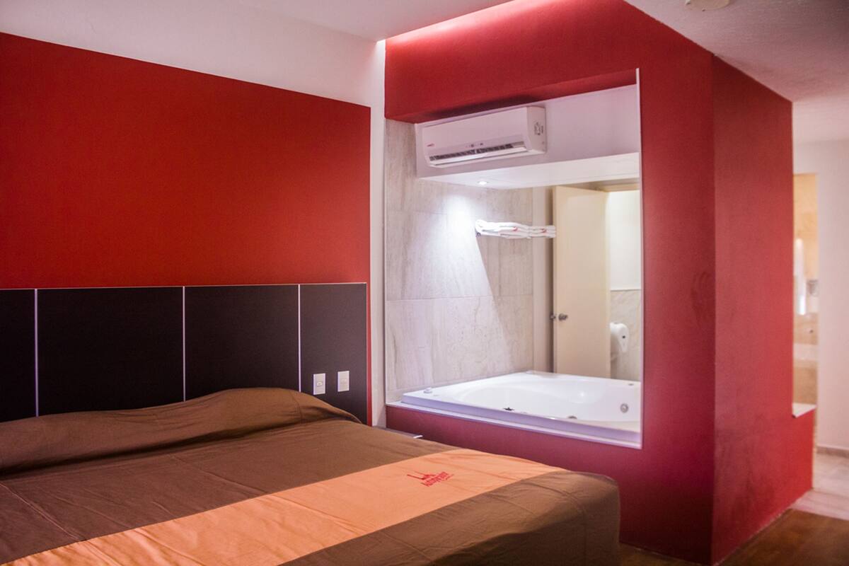 A modern bedroom with a large bed, a red accent wall, and a built-in window with a view of the bathroom.