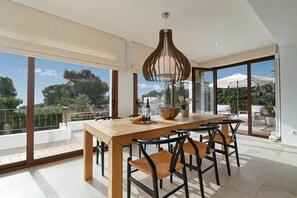 Zona pranzo con tavolo e sedie in legno, lampadario e ampie finestre che offrono vista su vegetazione e palazzi.