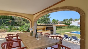 An outdoor patio with a wooden table and chairs, a barbecue area, and a pool in the background.