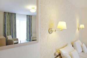 A hotel room with a bed, two wall-mounted lamps, a large mirror, a chair, and a view of the outside through curtains.