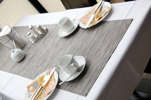 A neatly set dining table with white cups, saucers, and cutlery on a grey placemat.