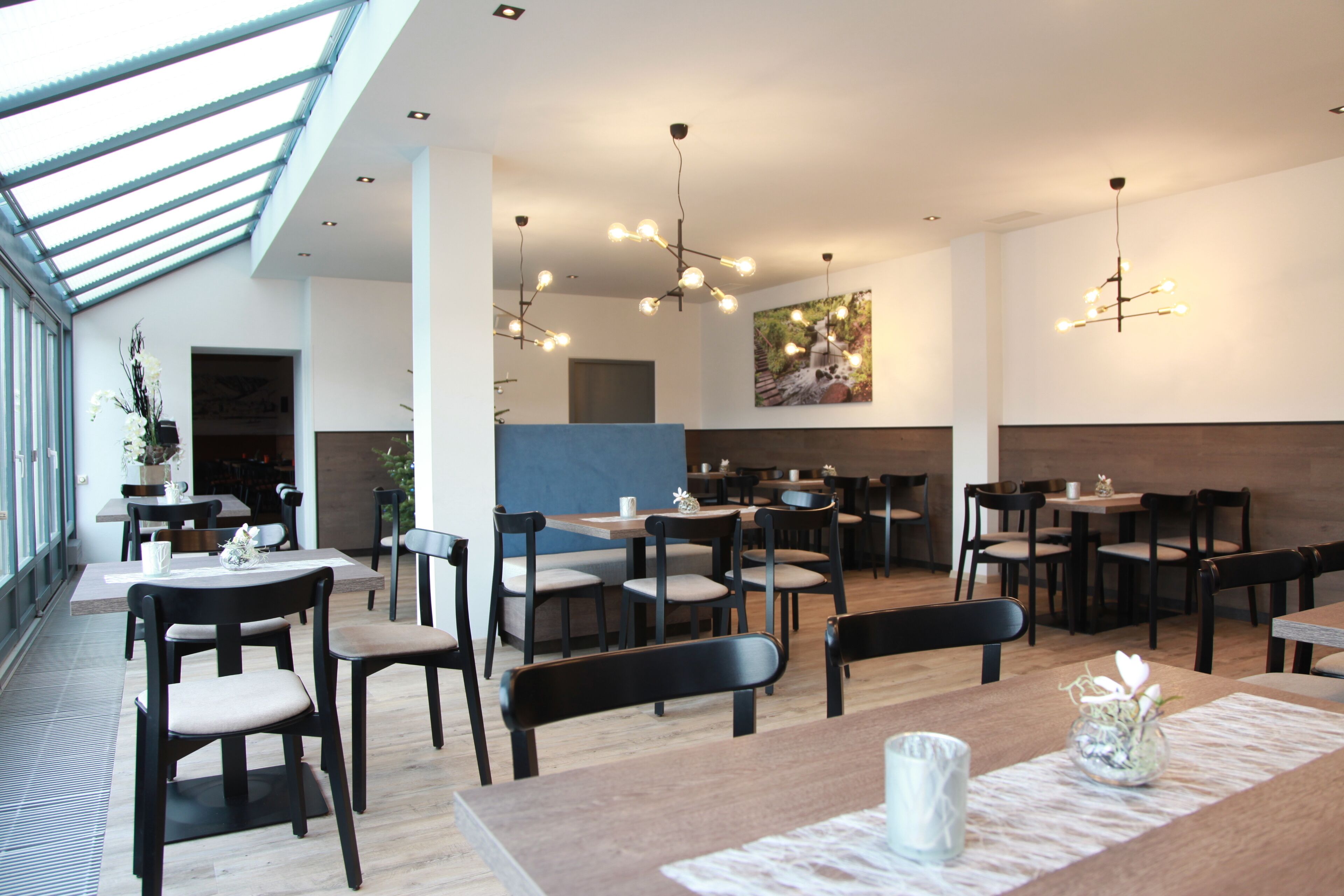 Un restaurante moderno con techo alto, luz natural que entra por tragaluces e iluminación contemporánea. Las mesas están dispuestas para comer, y hay sillas y una barra.