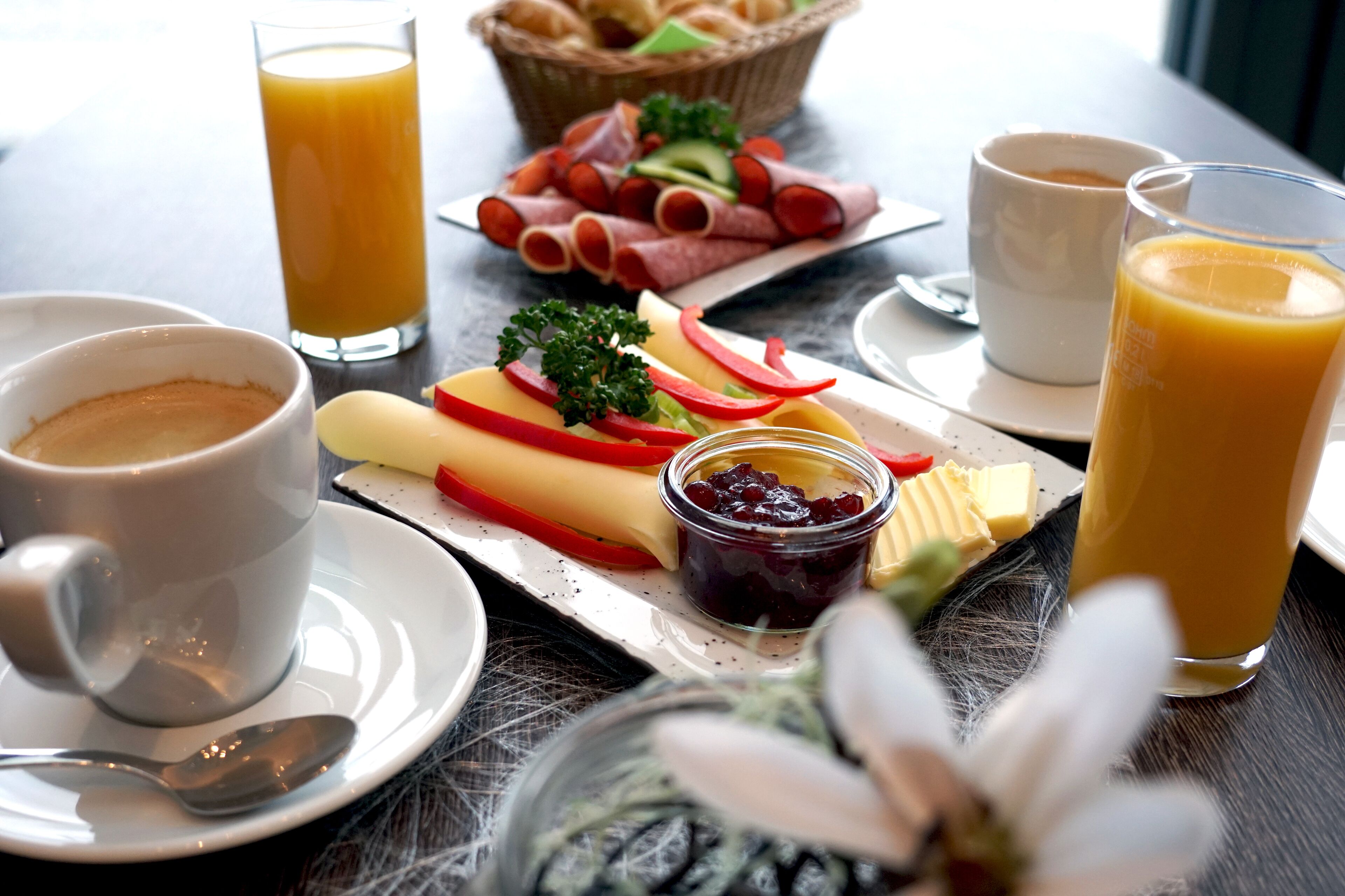 Una mesa de desayuno con un plato de jamón en rodajas, queso y verduras, acompañado de dos vasos de zumo de naranja y una taza de café.